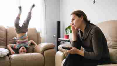 A mother sitting on a couch trying to actively ignore her child with ADHD who is doing a handstand on another couch -- an attention-seeking behavior.