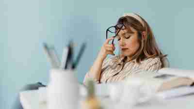 A woman, stressed and with closed eyes holds her glasses against her head. She thinks, "I don’t feel like an adult because of how ADHD impacts me, which I only discovered later in life."