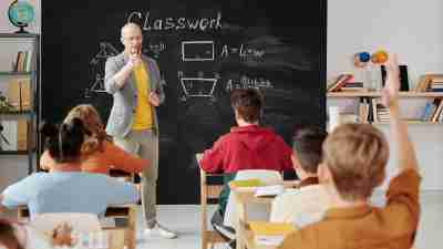 Students in a classroom facing the teacher. The teacher is pointing to a student in the back of the class who has his hand raised.