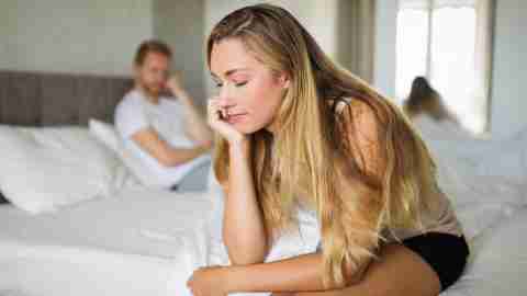 A woman is sitting on the edge of a bed, slouched over, with her head in her hand. A man can be seen in the background.